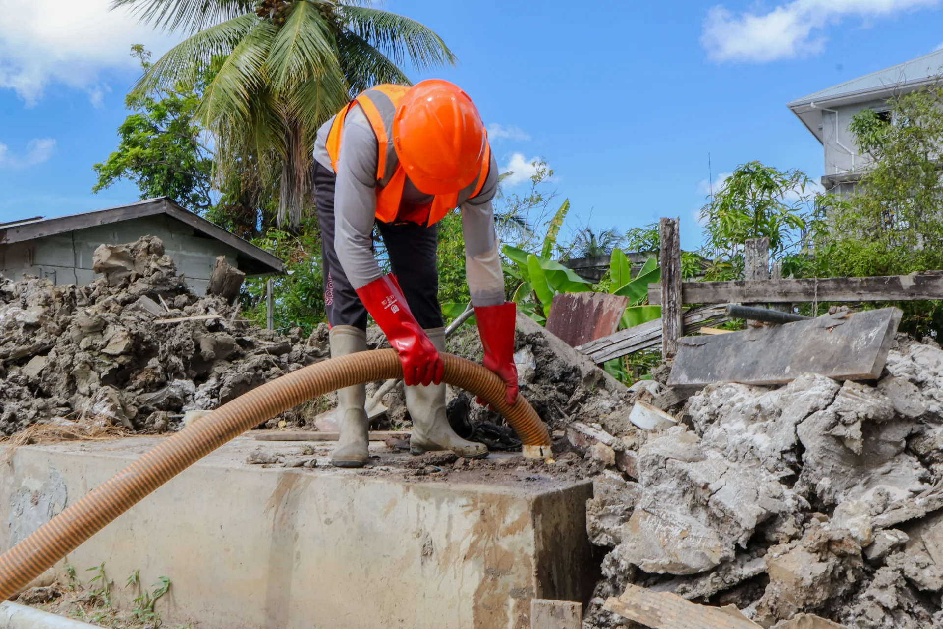 A CEVONS worker in an orange hard hat and high-visibility vest guiding a suction hose into an open concrete septic chamber on a cleared site.