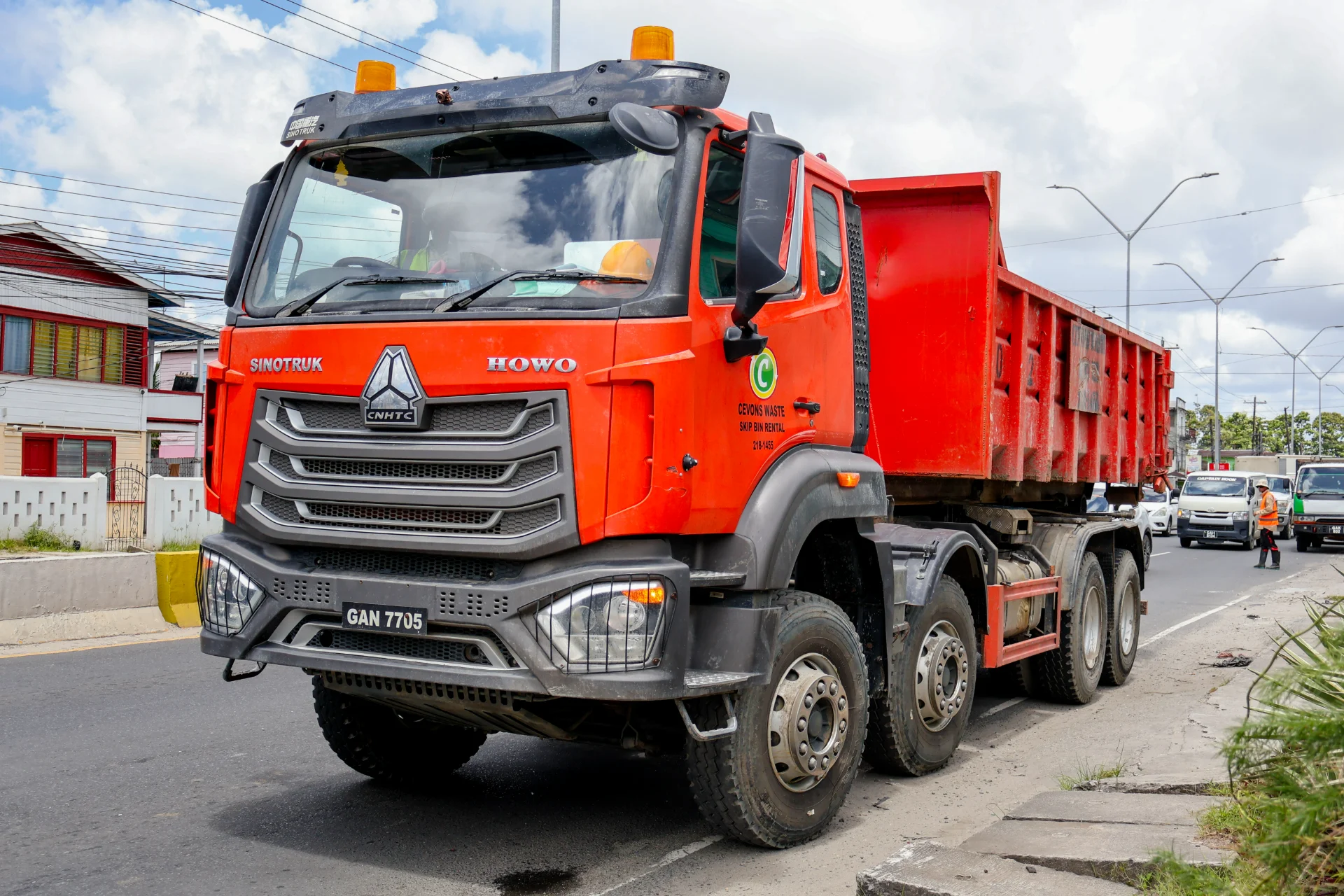 Orange CEVONS Sinotruk HOWO roll-off truck carrying a red skip bin on a Georgetown street.
