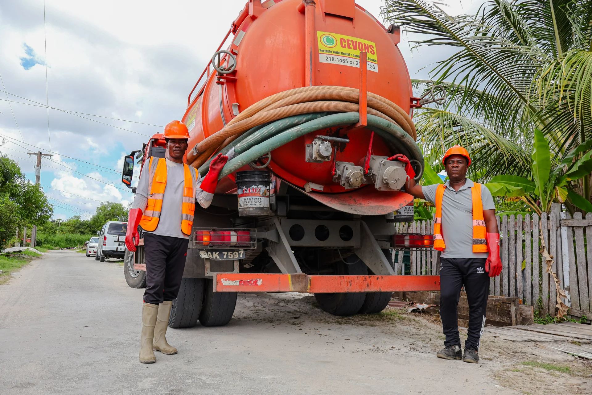 Two CEVONS crew members in orange hard hats, high-visibility vests and red gloves standing at the rear of an orange vacuum tanker with suction hoses coiled across the tank.